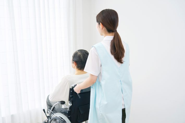 室内で高齢者女性の乗る車椅子を押す介護士　